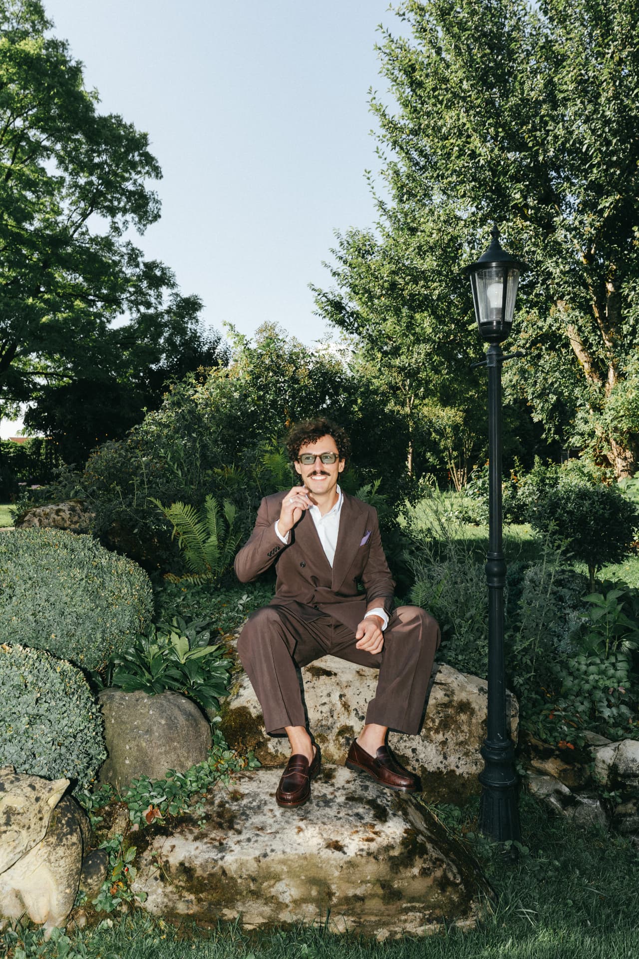 Bräutigam im braunen Anzug sitzt auf Stein im Garten – Portrait Hochzeitsfotograf Augsburg