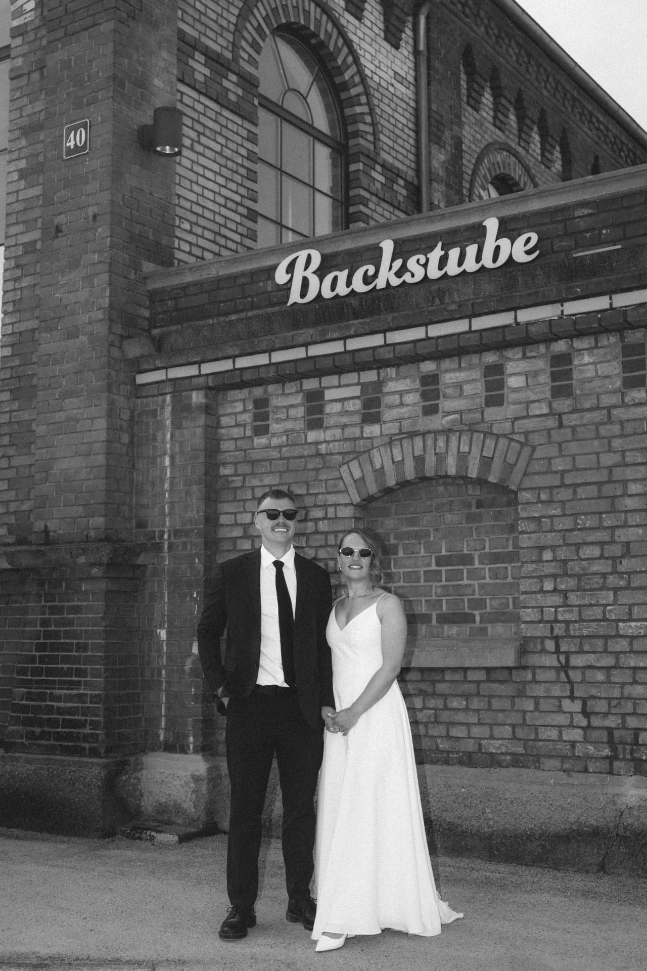 Brautpaar mit Sonnenbrillen vor der Backstube in Schwarzweiß – moderner Hochzeitsfotograf Augsburg