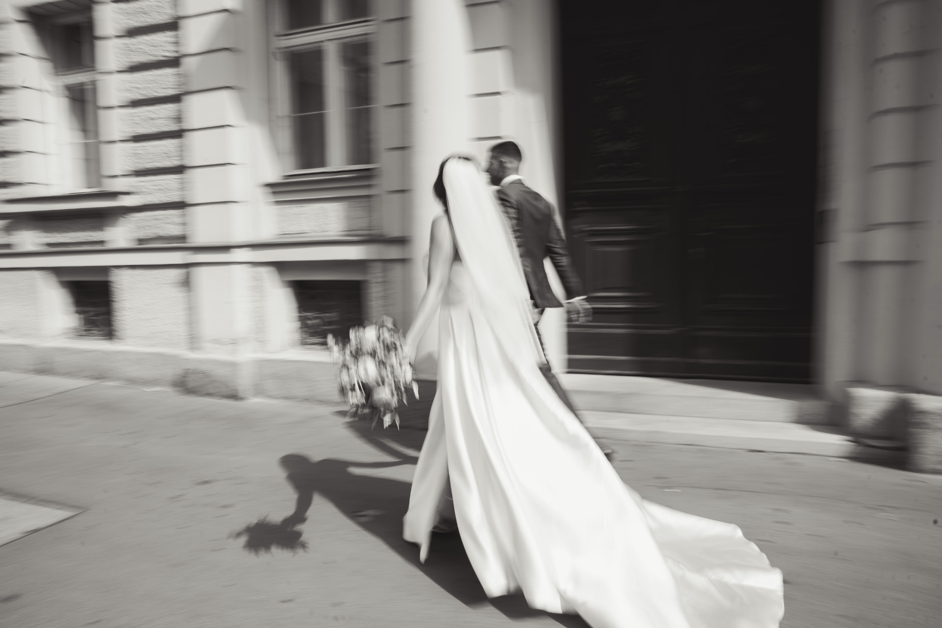 Schwarzweiß Aufnahme des Brautpaars mit langer Schleppe auf dem Weg zum Portal – Hochzeitsfotograf Augsburg